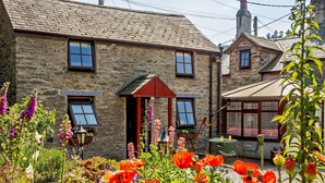 Un bâtiment en pierre à l’architecture traditionnelle, avec une porte d’entrée et un petit jardin agrémenté de différentes fleurs.