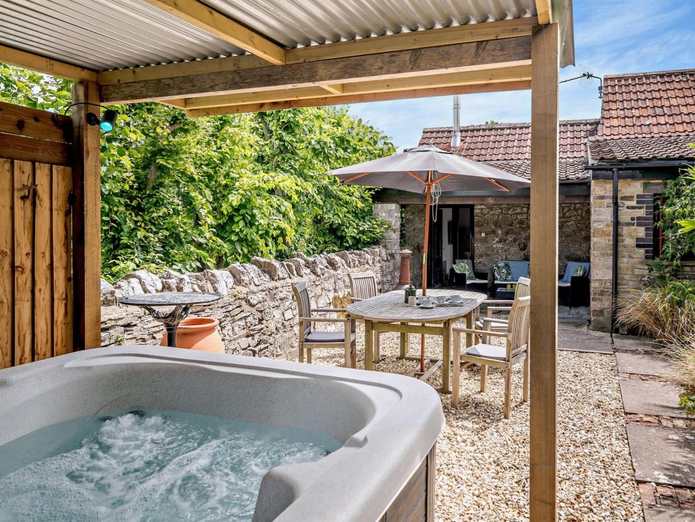 A hot tub under a wooden pergola with a stone wall and a table with chairs in the background.