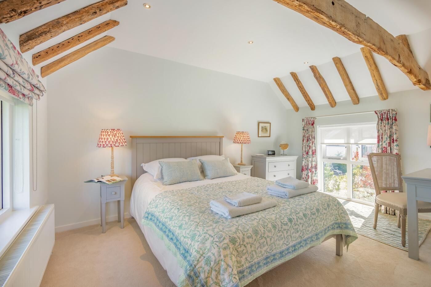 Quarto com cama, mesas de cabeceira, cômoda, janela com cortinas e vigas de madeira aparentes.