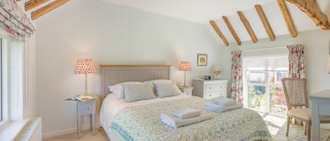 Un dormitorio con cama, mesitas de noche, cómoda, ventana con cortinas y vigas de madera a la vista.