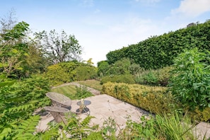 A garden patio with a wicker table and chairs, surrounded by lush greenery and a hedge. - 2 Bed in Cranbrook (oc-51229) (Goudhurst)