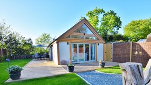 A modern house with a triangular roof, large sliding glass doors, a patio with outdoor furniture, and a well-maintained garden. - 1 Bed in Lymington (oc-50723) (Sway)