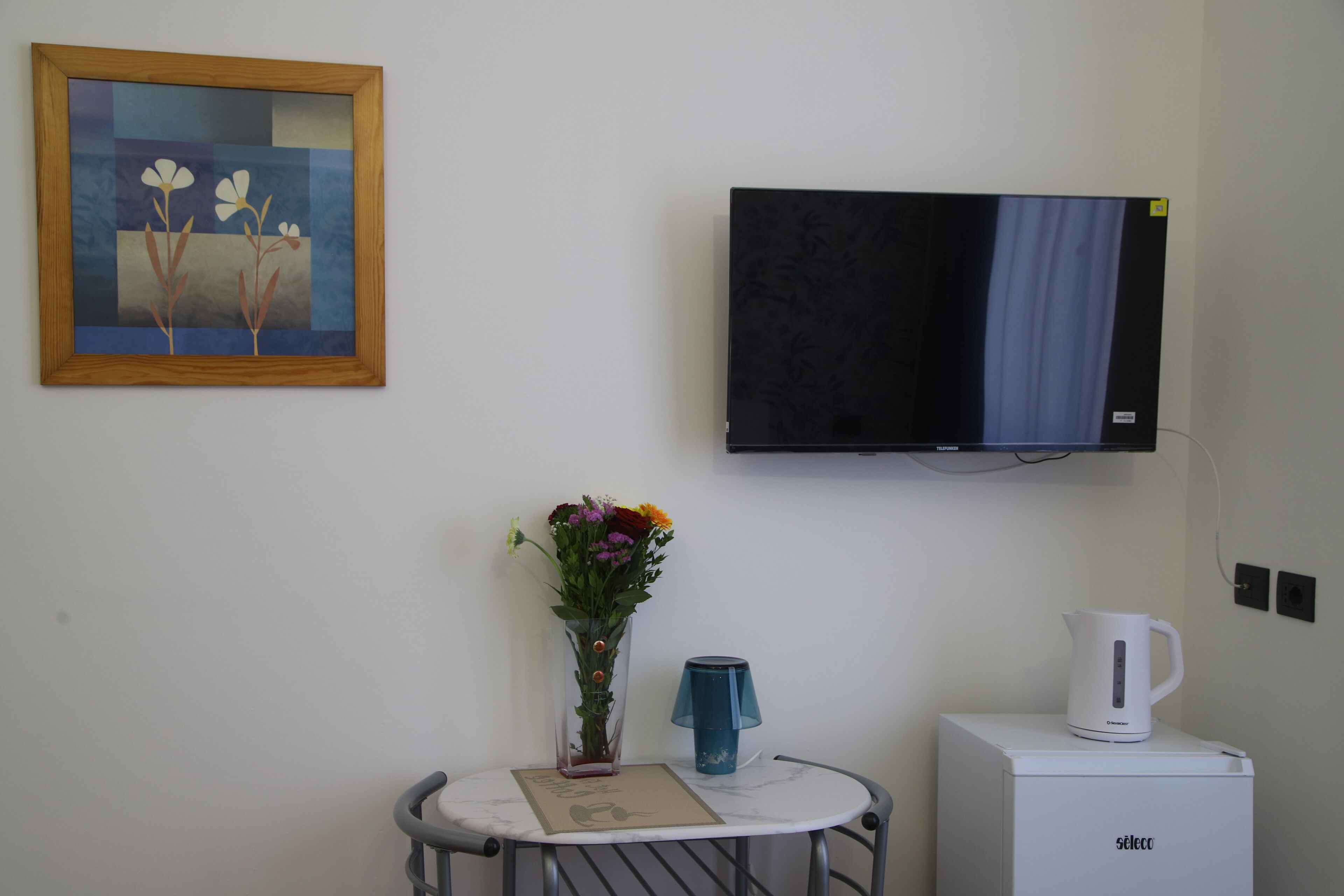 A room with a TV mounted on the wall, a small table with a vase of flowers, a kettle, and a framed picture.