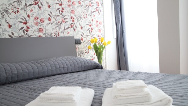 A neatly made bed with grey bedding, a floral wallpaper, and a vase with yellow flowers. - LA TORRETTA (Savona)