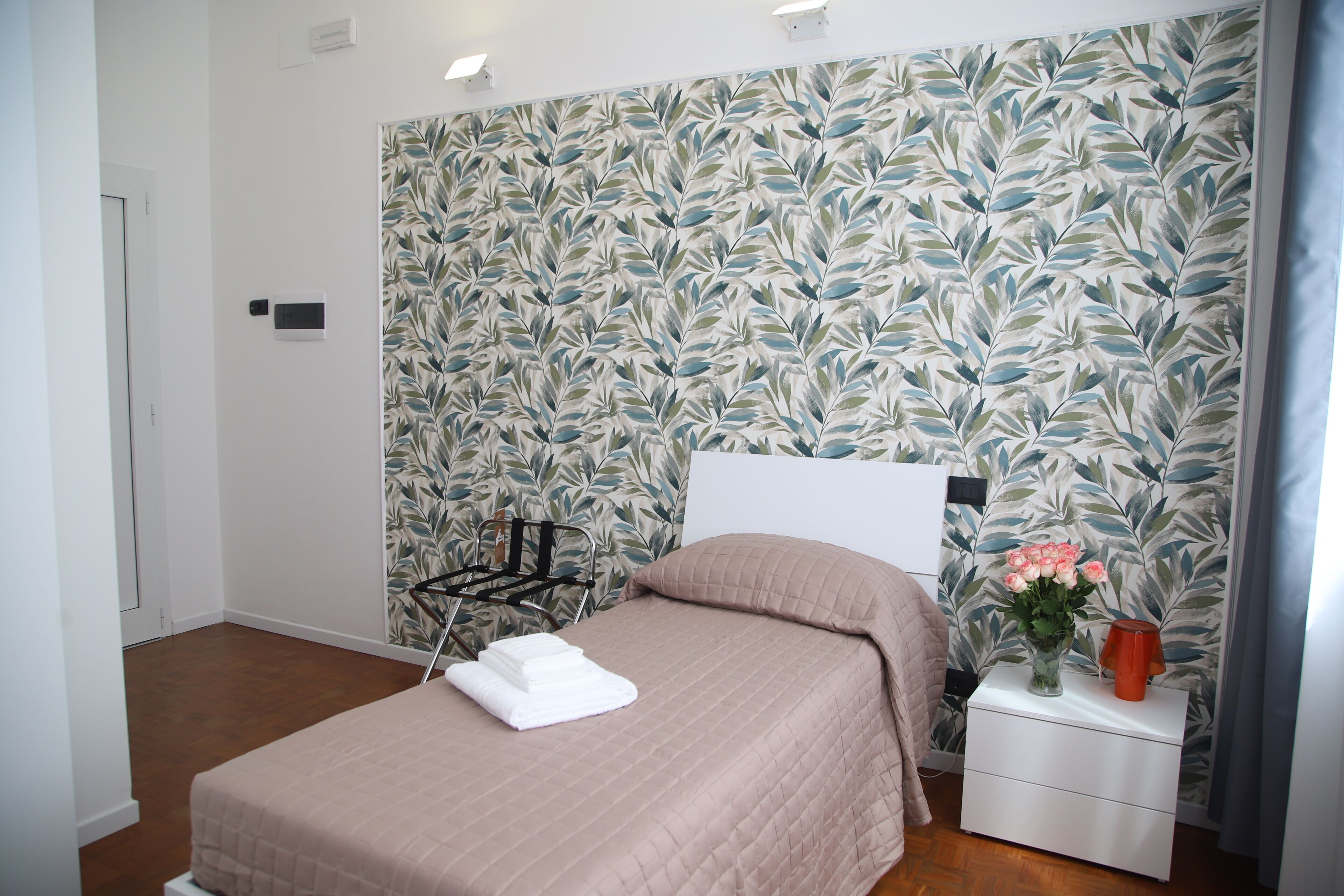 Ein ordentlich bezogenes Bett mit einer rosafarbenen Tagesdecke, ein weißer Nachttisch mit einer Blumenvase und eine Wand mit einem Blattmuster.
