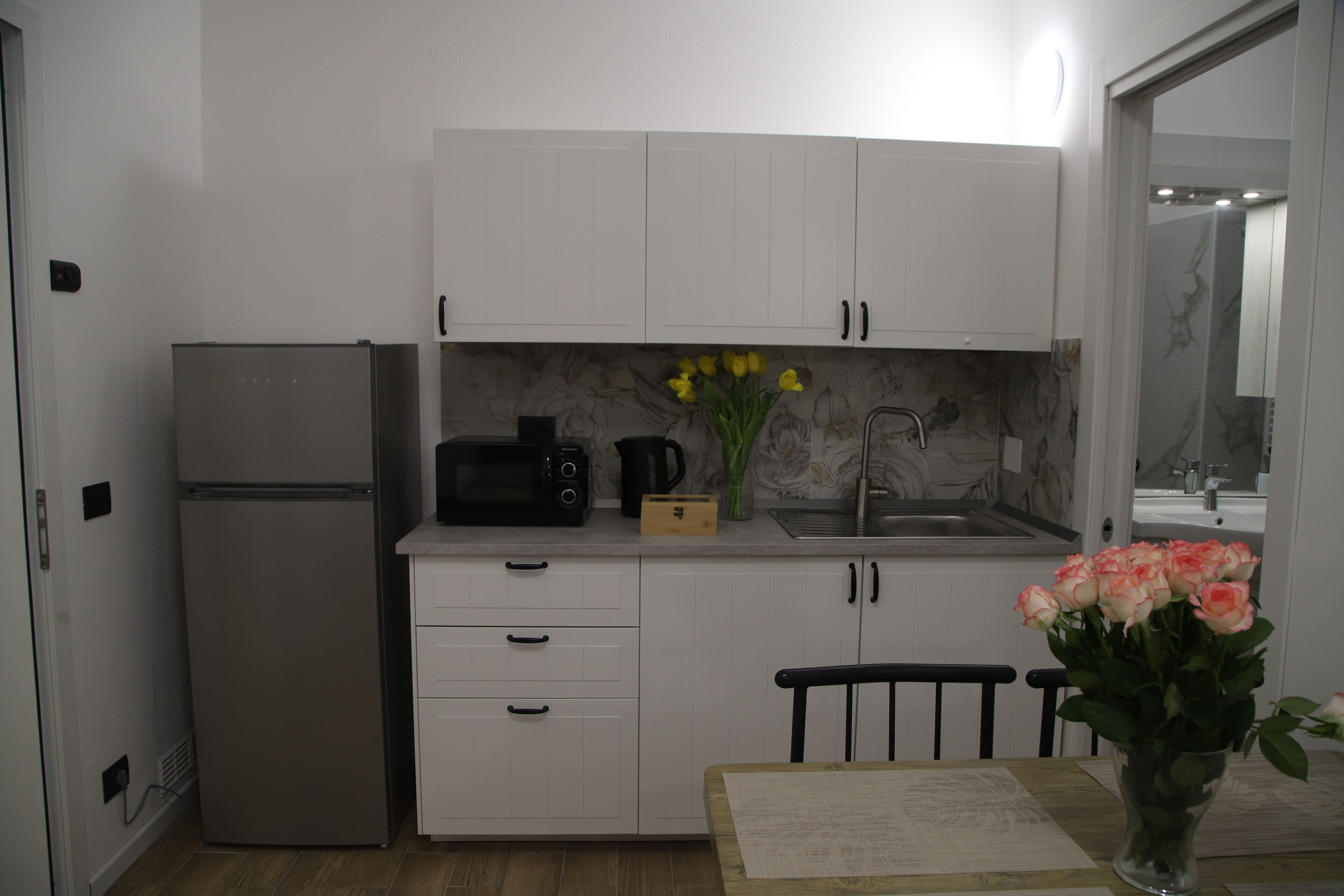 A compact kitchen with white cabinets, a refrigerator, a microwave, a coffee maker, and a vase with flowers.