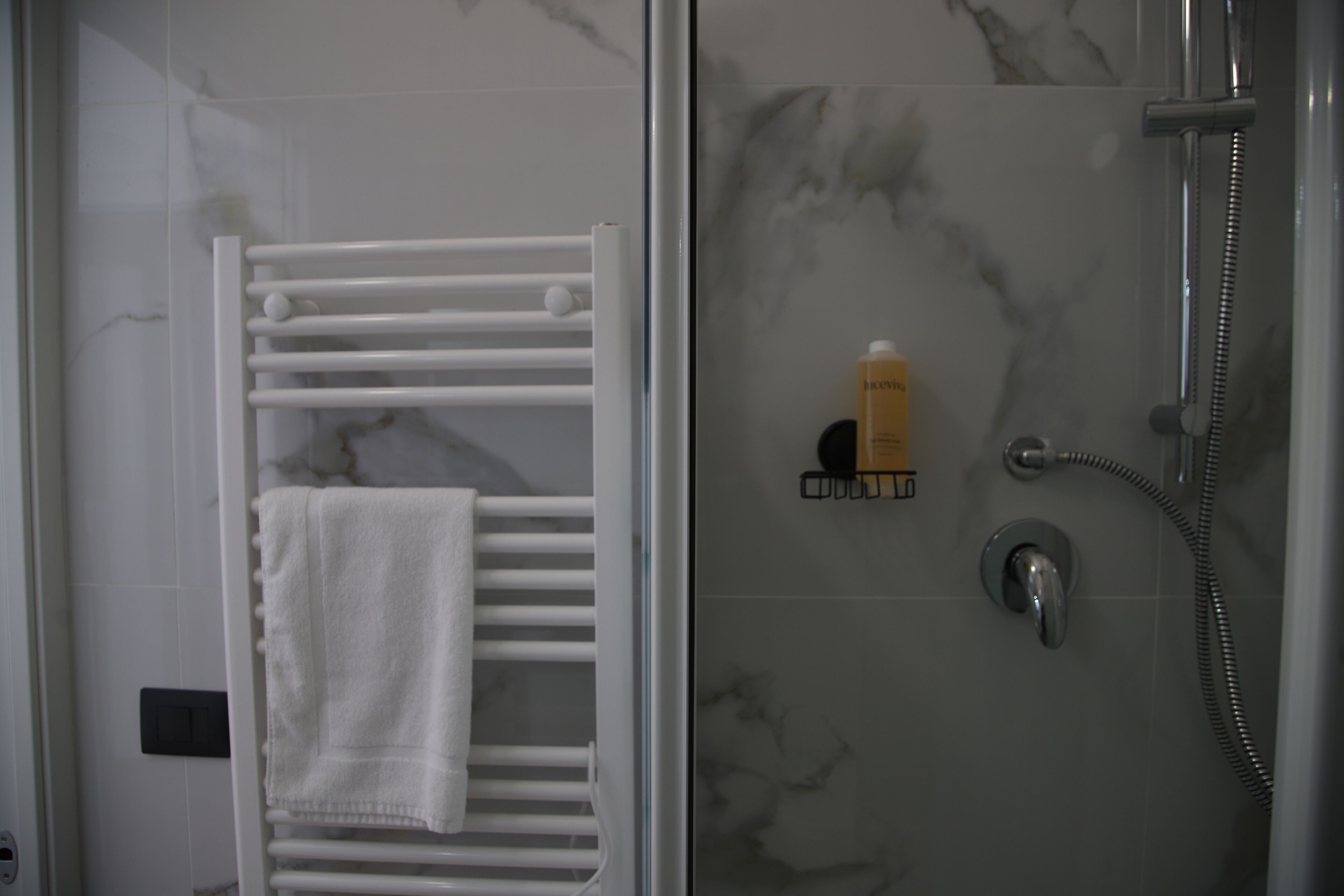 Kylpyhuone, jossa on lasiseinällä varustettu suihku, valkoinen pyyhekoukku ja pullo shampoota.