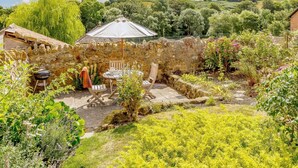 Eine rustikale Steinmauer, ein Holztisch mit Stühlen und ein Garten mit verschiedenen Pflanzen und Blumen.