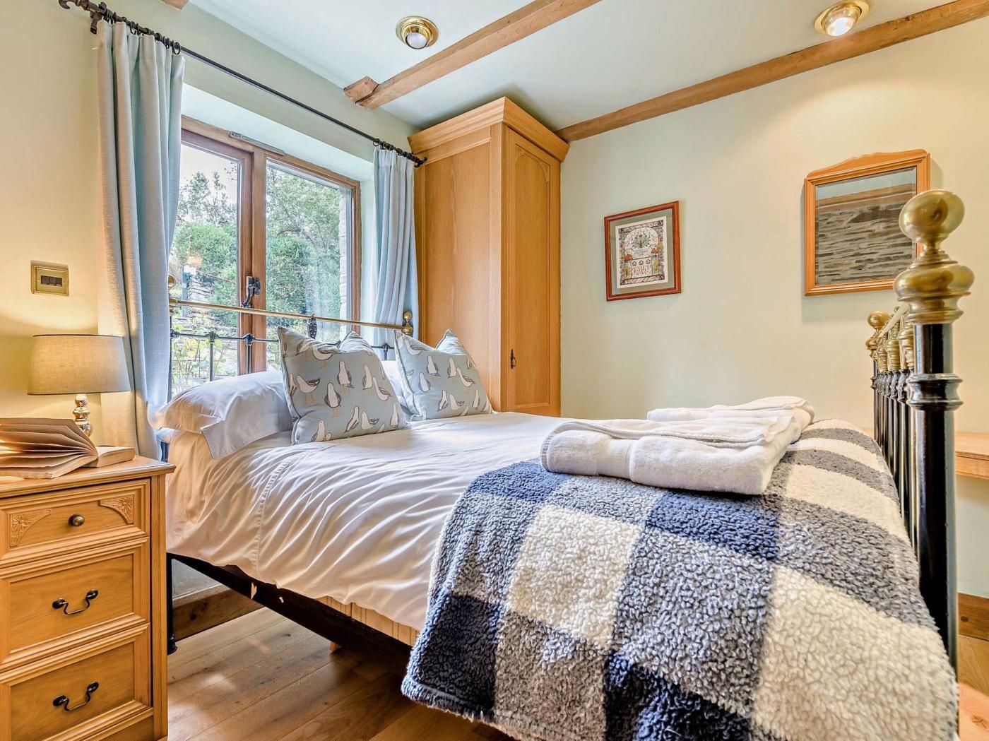 A bedroom with a bed, wooden dresser, window with curtains, and a view of greenery outside.