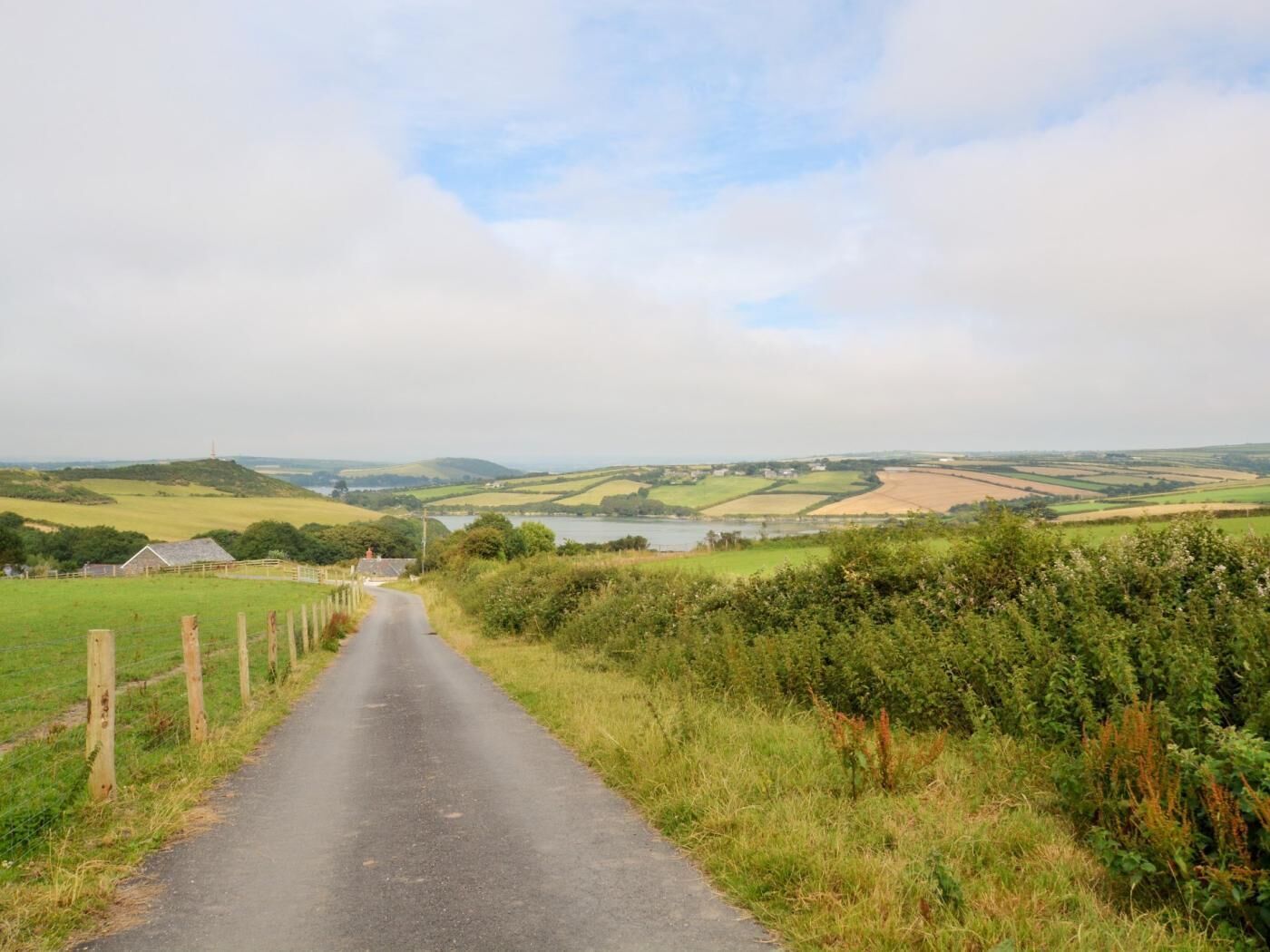 A rural road leading through a countryside with fields and a body of water in the distance.