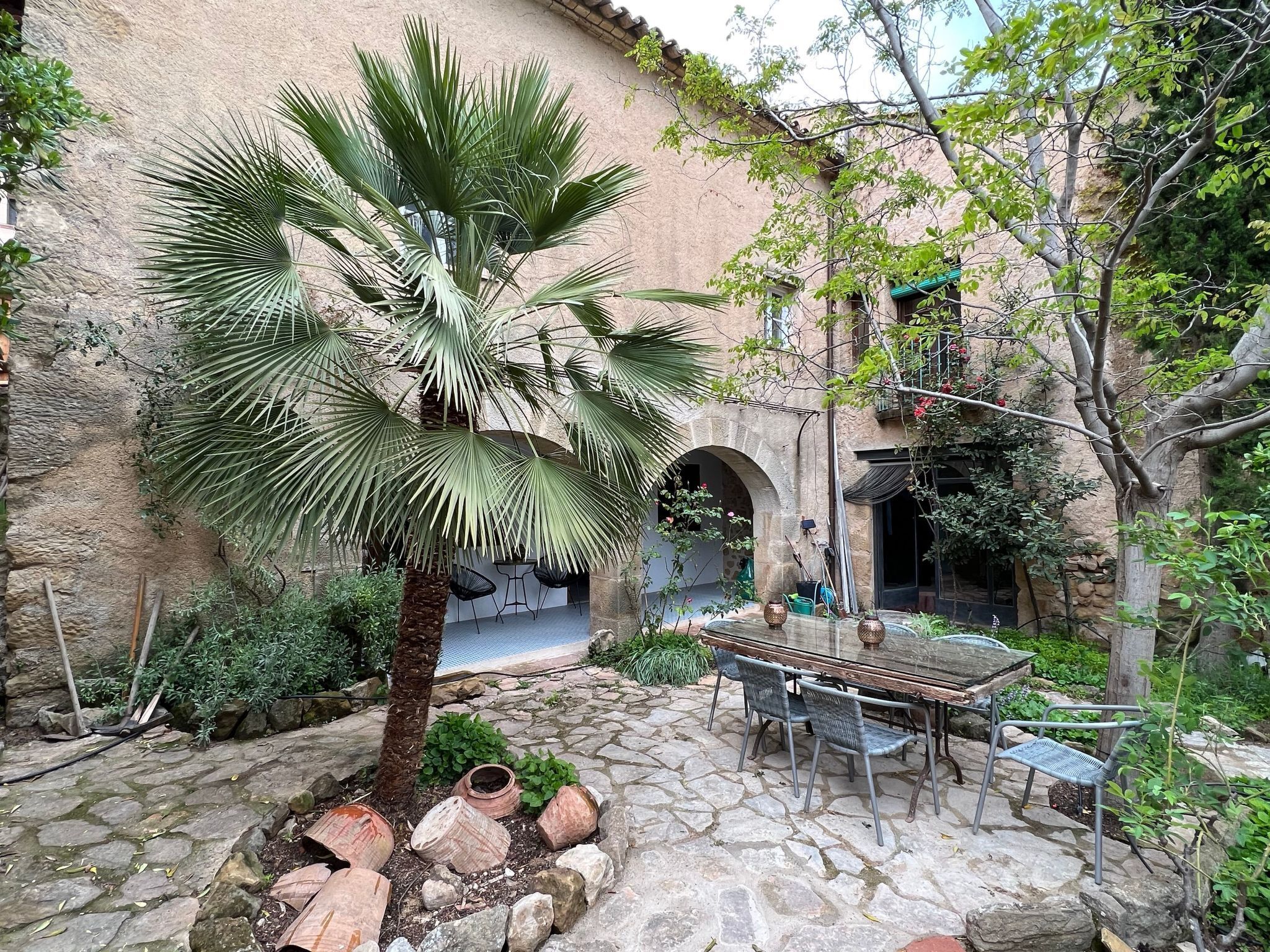 A courtyard with a palm tree, stone flooring, a table with chairs, and a building with arches.
