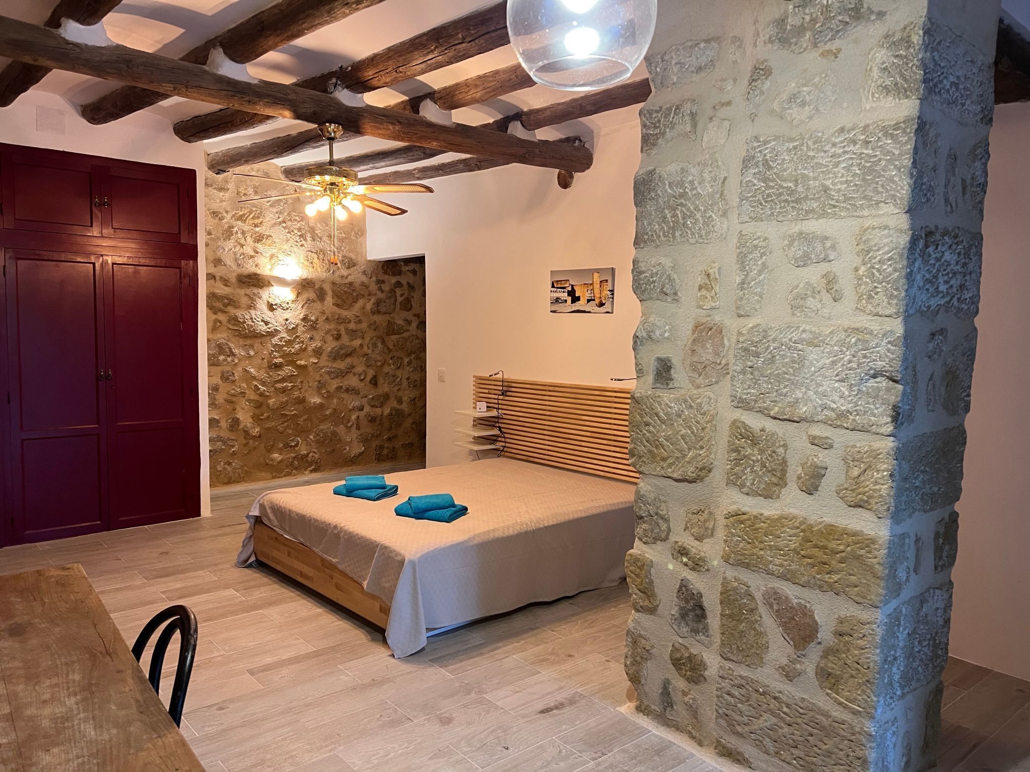 A bedroom with a stone wall, wooden ceiling beams, a bed with a wooden headboard, and a red door.