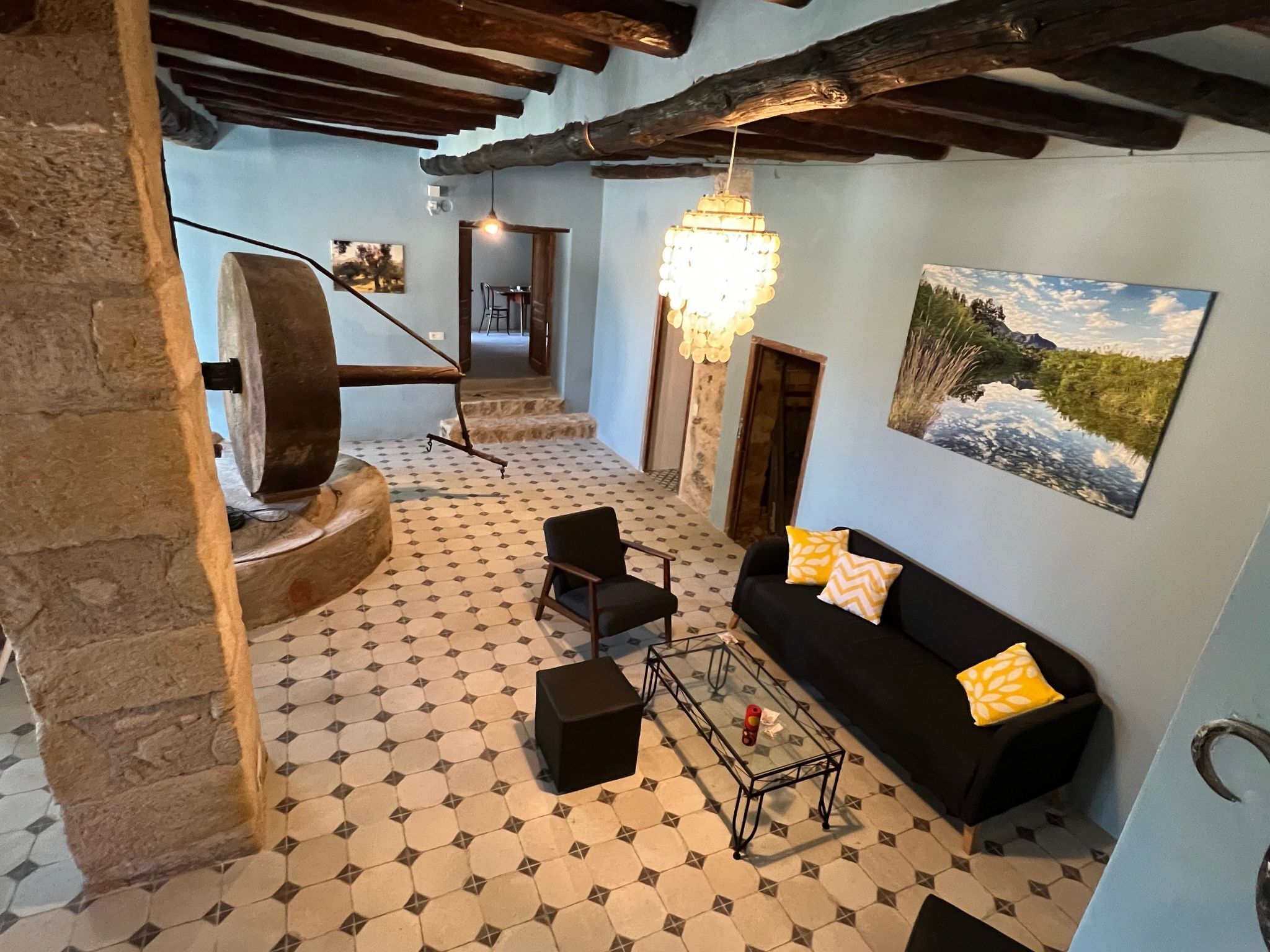 Una habitación con pared de piedra, un sofá negro con almohadones amarillos, una mesa de centro de vidrio, un sillón negro, una lámpara colgante y un cuadro de un paisaje.