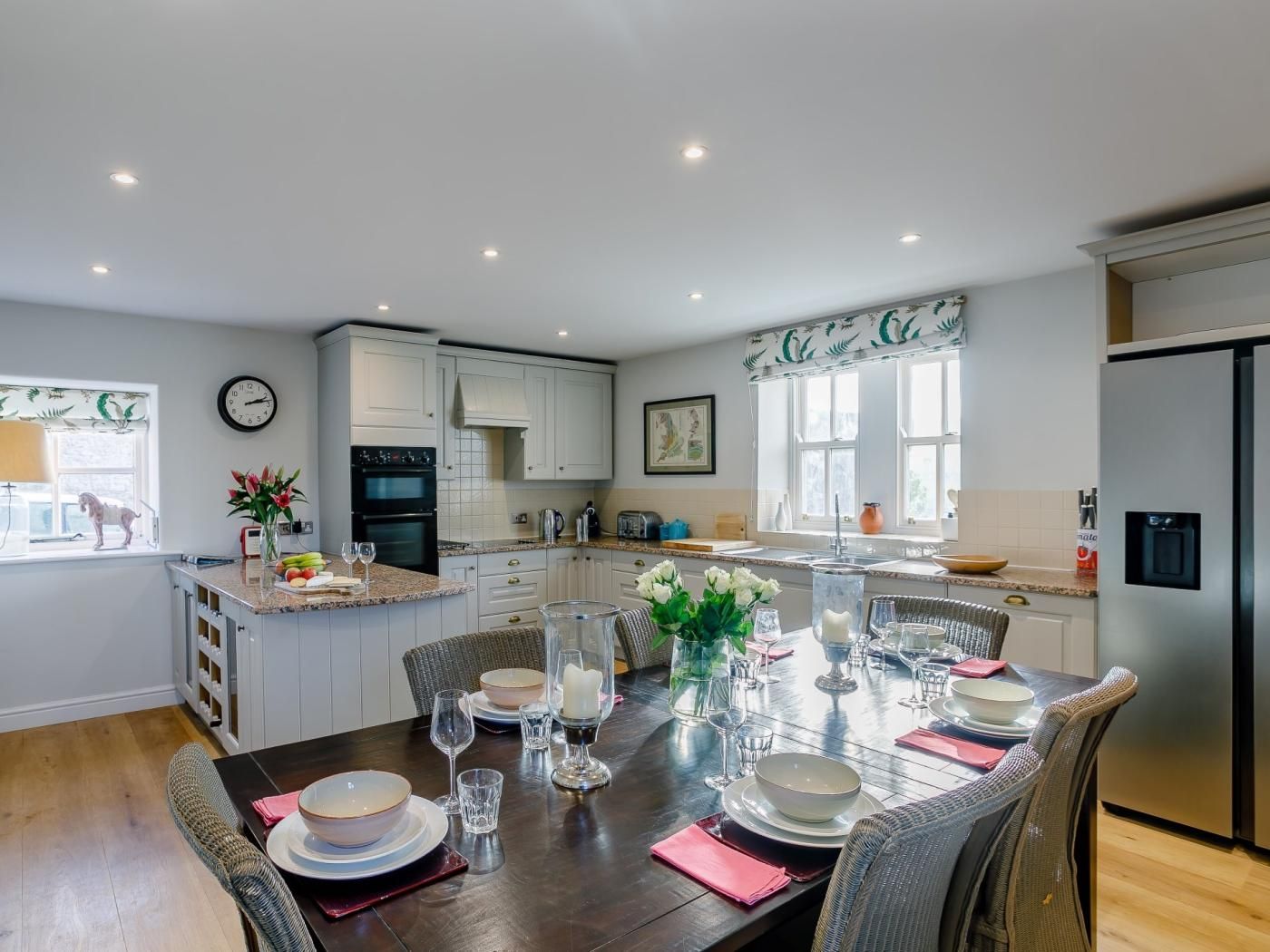 Une cuisine moderne avec des meubles blancs, une table à manger dressée pour six personnes et un grand réfrigérateur.