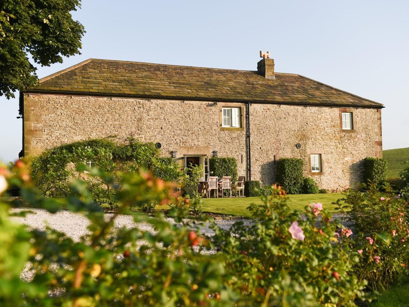 Un bâtiment en pierre avec un toit à deux versants, une cheminée et un petit jardin comprenant une pelouse et des arbustes.