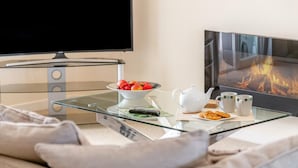 A modern living room with a glass coffee table, a bowl of fruit, a teapot, and two mugs. A flat-screen TV is mounted on the wall, and a fireplace is visible in the background. - 3 Bed in Bideford (oc-51790) (Hartland)