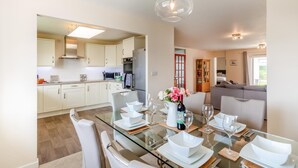 A modern kitchen with white cabinets, a dining area with a glass table, and a living room with a sofa. - 3 Bed in Bideford (oc-51790) (Hartland)