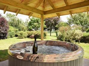Uma banheira de hidromassagem com pérgola de madeira, rodeada por um jardim com árvores e um banco.