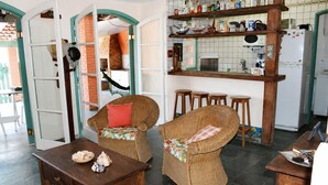 A cozy interior with wicker furniture, a wooden table, and a kitchen area with shelves and stools. - Family home in Lagoinha: sea view, barbecue and 24-hour security. (Ubatuba)