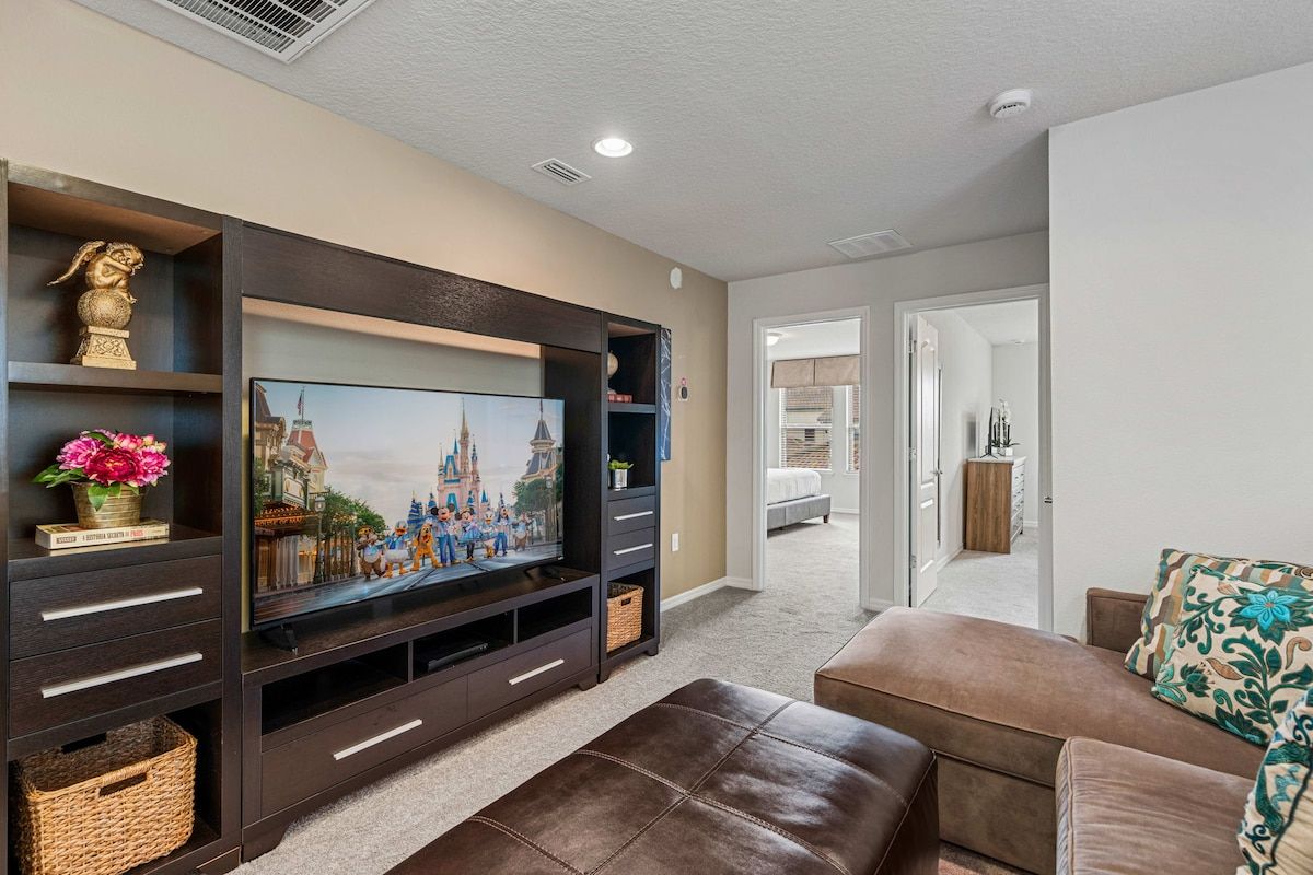 A living room with a flat-screen TV, dark wooden entertainment unit, brown leather sofa, and a hallway leading to a bedroom.
