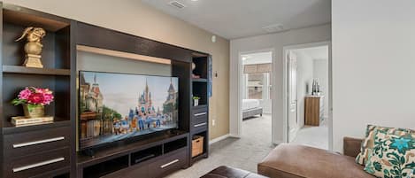 A living room with a flat-screen TV, dark wooden entertainment unit, brown leather sofa, and a hallway leading to a bedroom.
