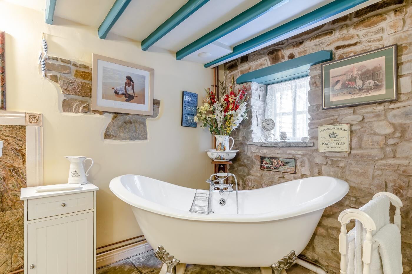A bathroom with a freestanding bathtub, a white sink, a mirror, a towel rack, and a stone wall with framed pictures and a clock.