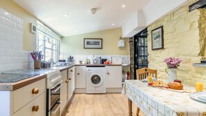 A compact kitchen with a washing machine, a dining area with a table set for breakfast, and a stone wall. - 1 Bed in Bude (oc-51546) (Gooseham)