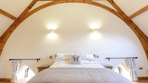 Une chambre à coucher avec un plafond voûté, un lit recouvert d’une courtepointe blanche, deux lampes de chevet et une fenêtre avec des rideaux.