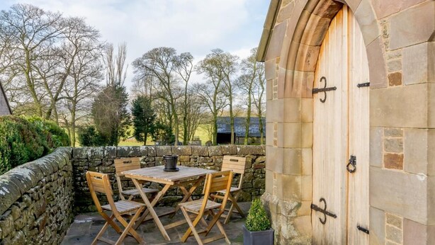 Un bâtiment en pierre avec une porte en bois, un mur en pierre et une table avec des chaises à l’extérieur.