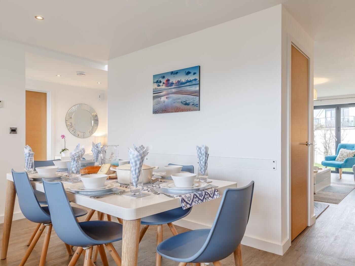Un espace repas moderne avec une table blanche dressée pour quatre personnes, des chaises bleues, un grand miroir et un tableau représentant un paysage.
