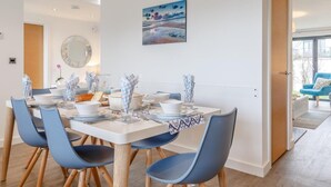 Et moderne spiseområde med et hvidt bord dækket til fire personer, blå stole, et stort spejl og et landskabsmaleri.