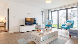 En moderne stue med et fladskærms TV, et glasbord, to blå lænestole og en stor skydedør, der fører ud til en have.