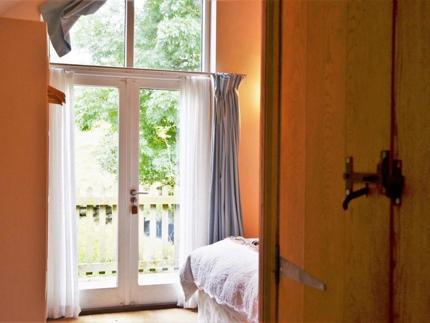 A room with a sliding glass door leading to a balcony, a bed with a white bedspread, and a wooden door with a metal handle.