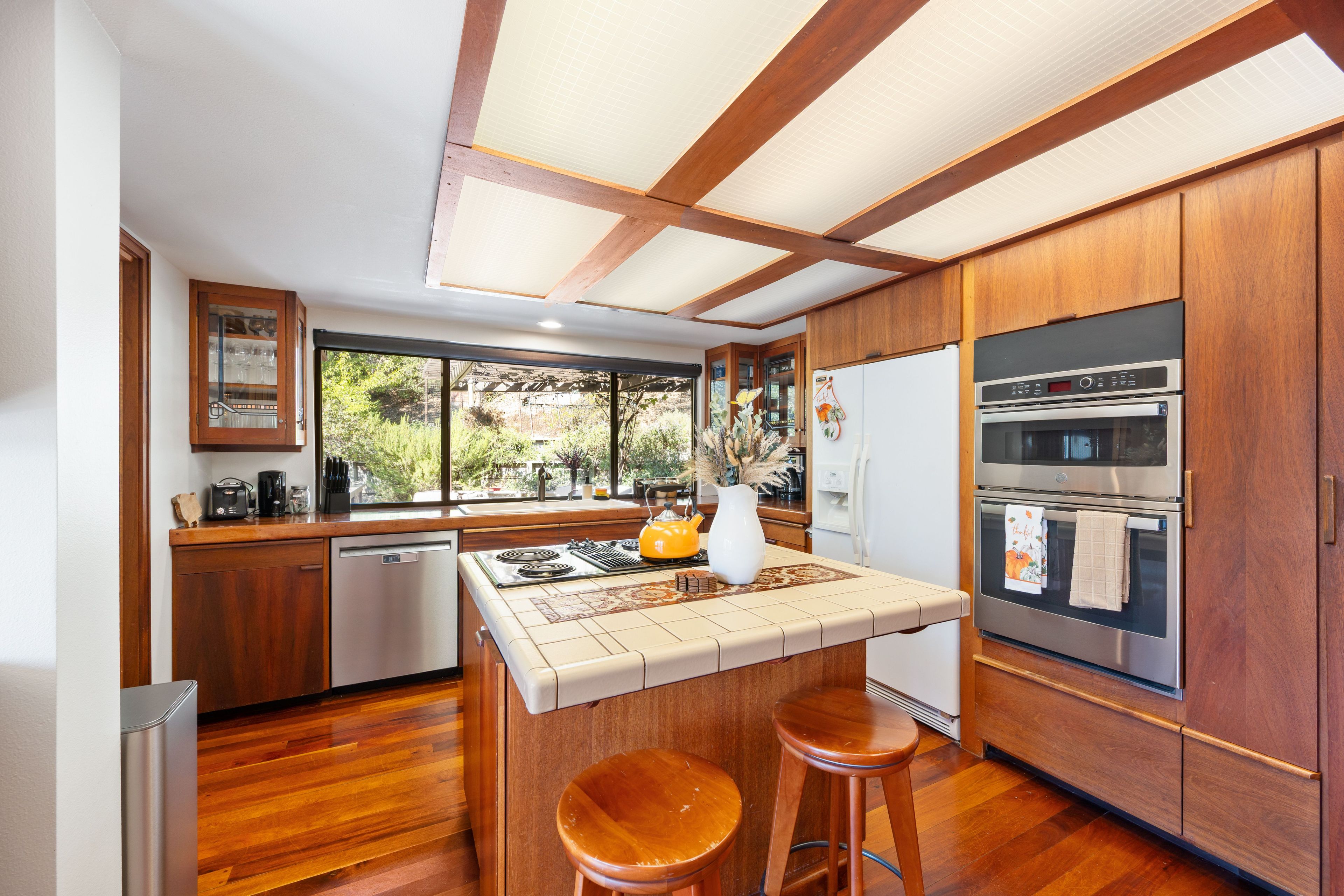 A modern kitchen with wooden cabinets, a built-in oven, and a large window overlooking greenery.