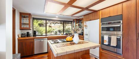A modern kitchen with wooden cabinets, a built-in oven, and a large window overlooking greenery.