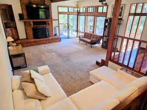A spacious living area with a fireplace, a television mounted on a brick wall, and a large window providing a view of the outdoors.