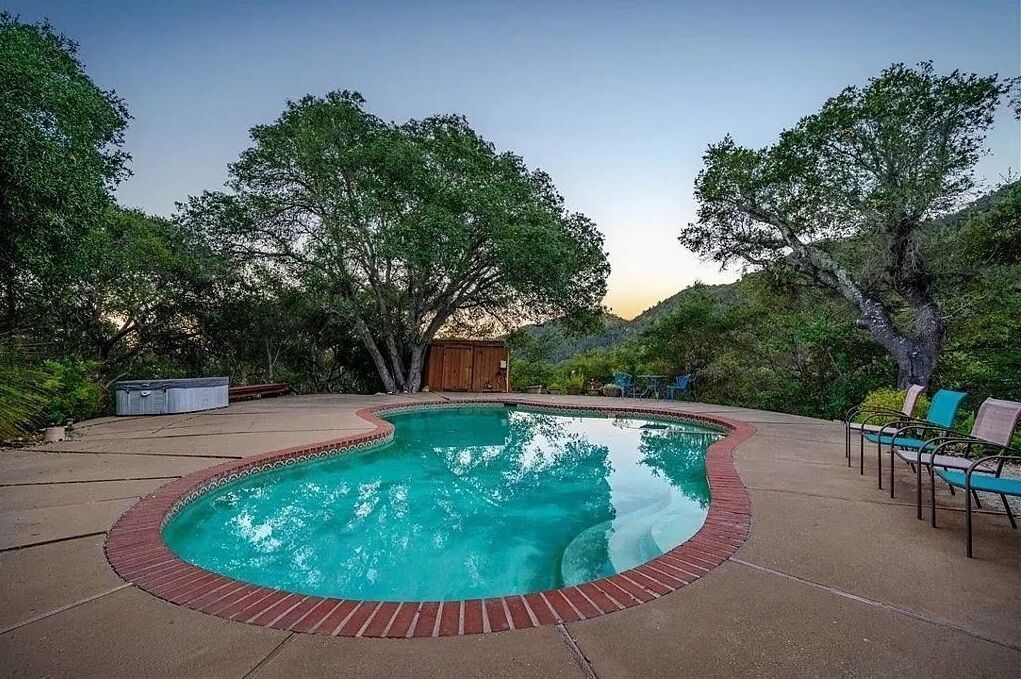 A swimming pool with a brick border, surrounded by trees and outdoor furniture.