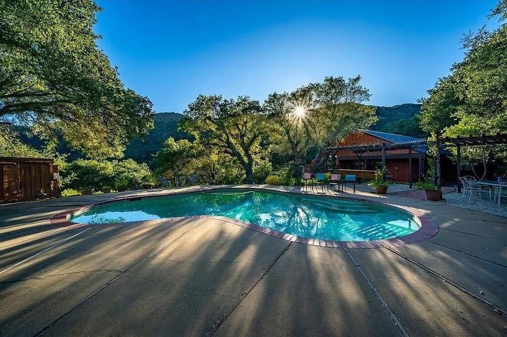 Une piscine avec une terrasse en bois, entourée d’arbres et bordée d’un bâtiment doté d’un toit en pente.