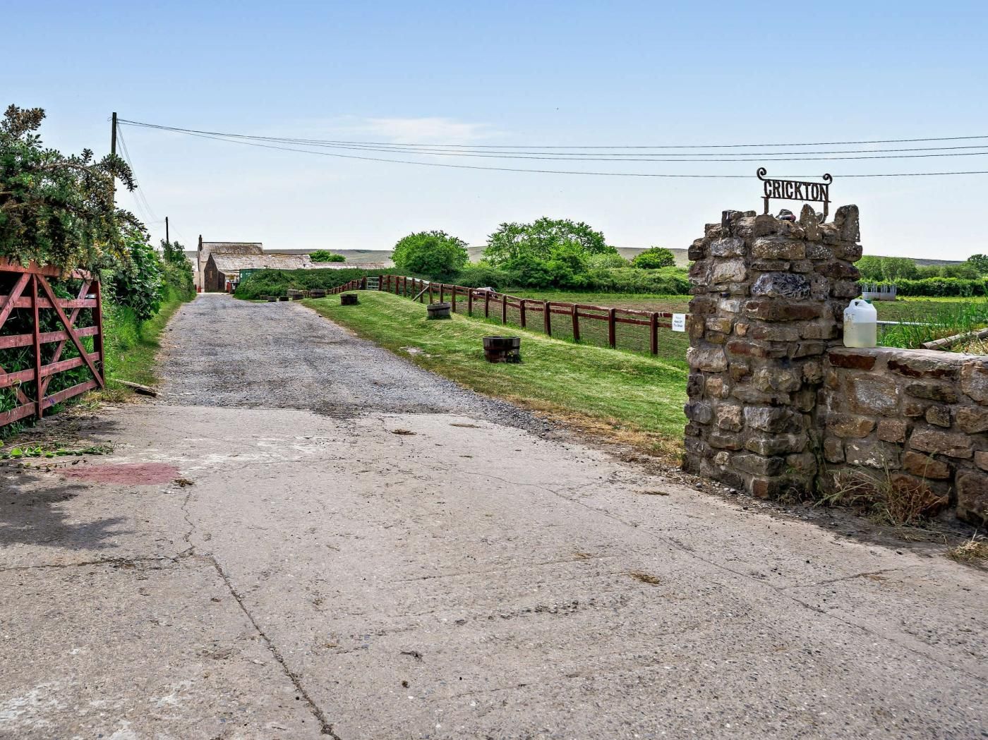 Een landelijke opgang met een stenen poort waarop 'CRICKTON' staat, een grindpad dat naar een gebouw leidt, en een omheind gebied met groen.