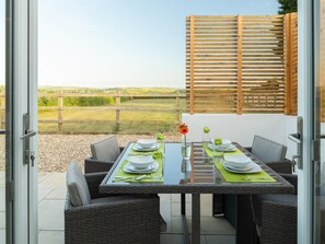 Un espace extérieur aménagé pour les repas, avec une table dressée pour quatre personnes, entourée de chaises en osier, d’une clôture en bois et offrant une vue sur la campagne.