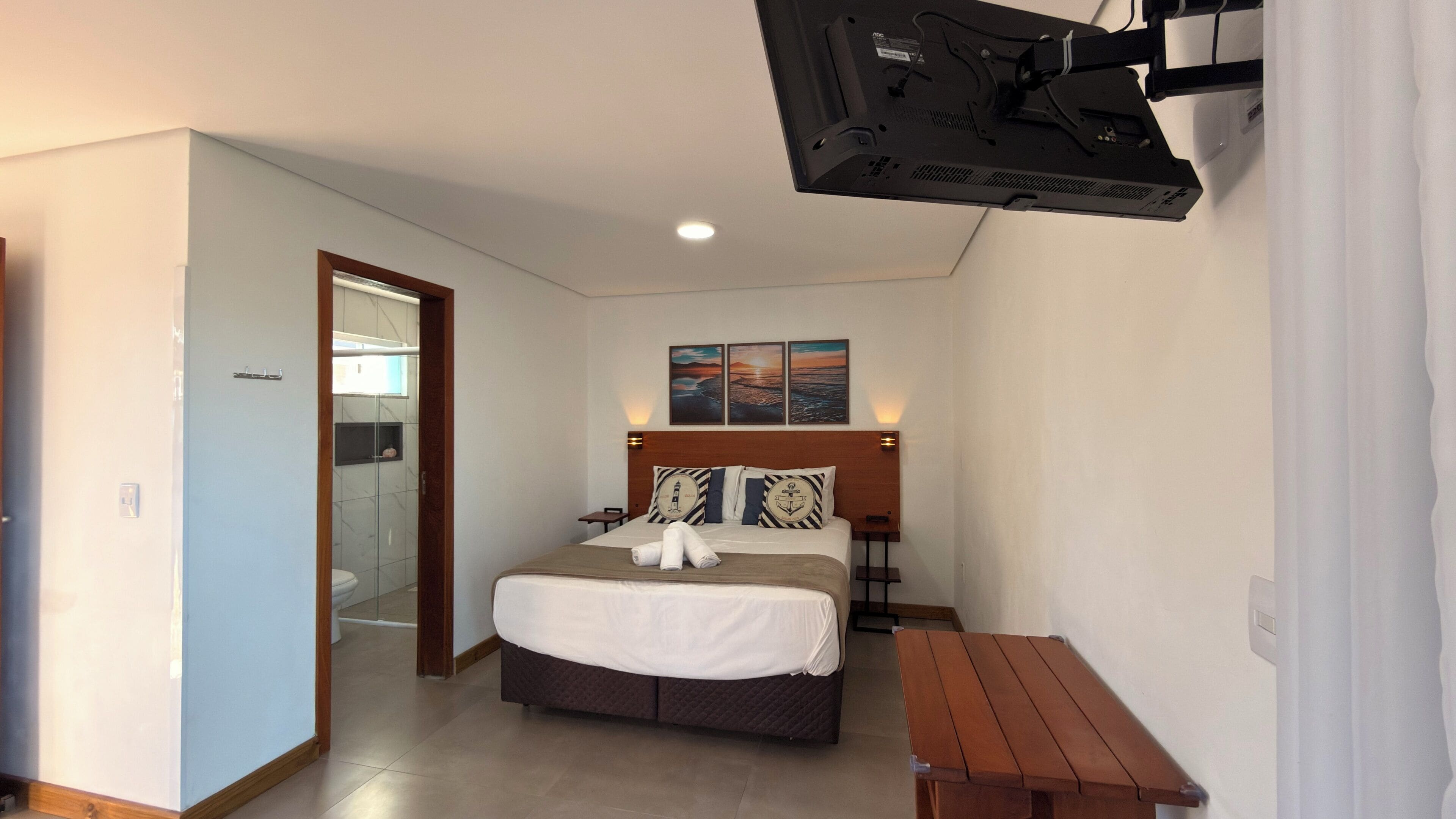 Habitación de hotel con cama, mesitas de noche, un banco de madera, zona de ducha y televisor montado en la pared.