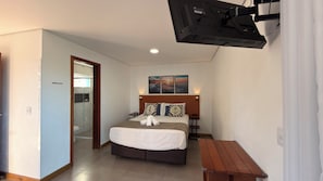 A hotel room with a bed, bedside tables, a wooden bench, a shower area, and a wall-mounted TV.