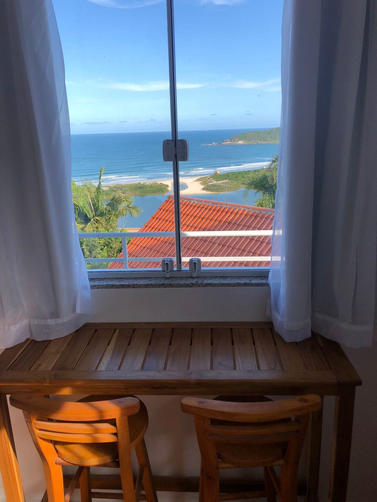 Una mesa de madera con dos sillas, una ventana con cortinas blancas y vistas al océano y un edificio con techo rojo.