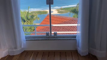 Una mesa de madera con dos sillas, una ventana con cortinas blancas y vistas al océano y un edificio con techo rojo.