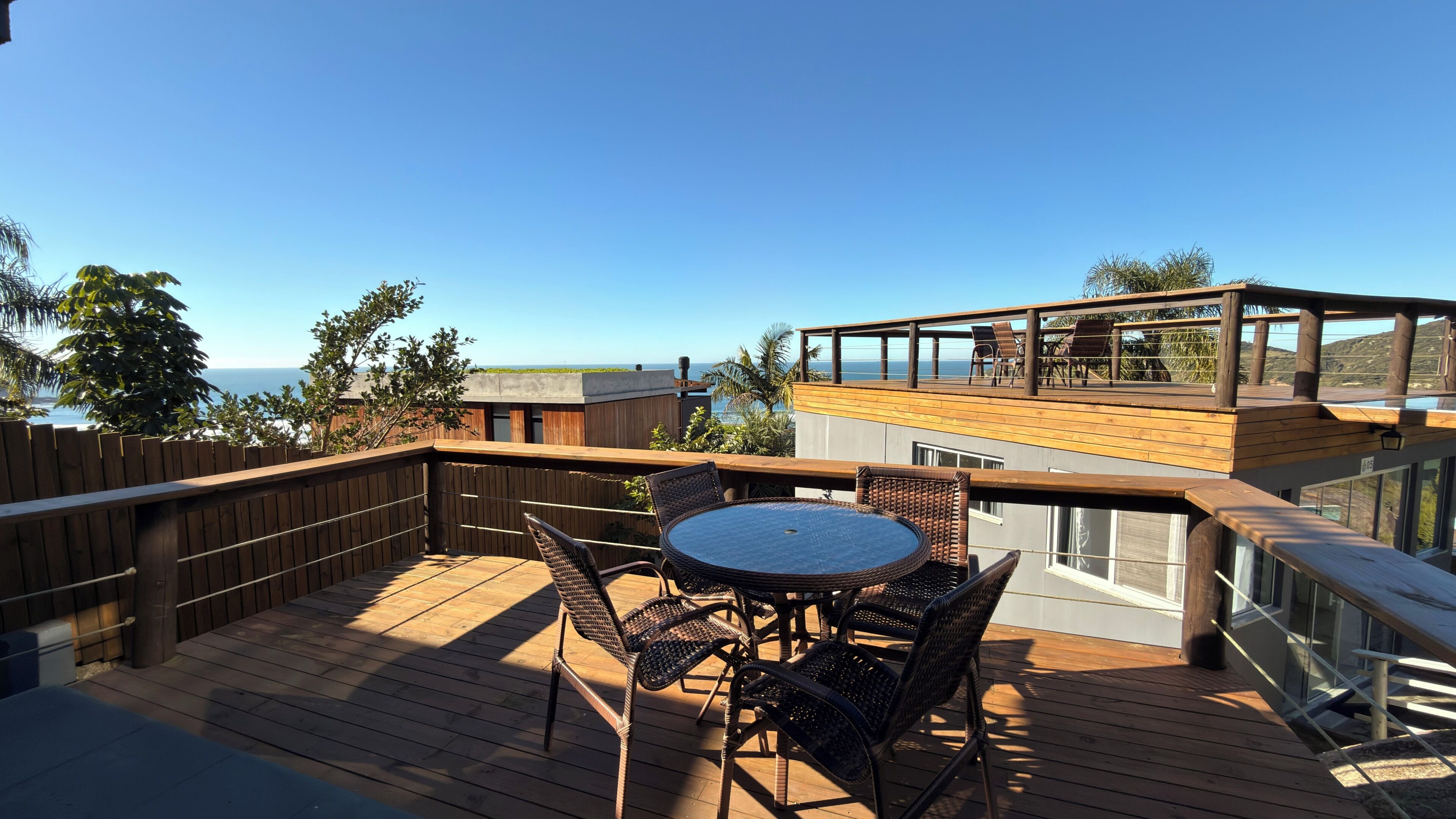 Una terraza de madera con una mesa redonda y sillas, con vista a un cuerpo de agua y colinas.