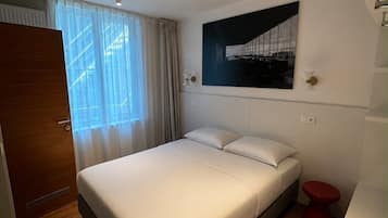 Ein modernes Hotelzimmer mit einem großen Bett, einem roten Akzentstuhl, einem Wandbild und einem Fenster mit transparenten Vorhängen.