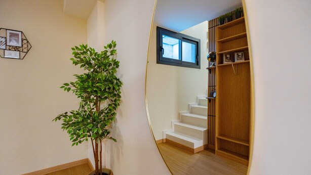 Un couloir avec un pot de fleurs, un miroir qui reflète un escalier et une fenêtre.