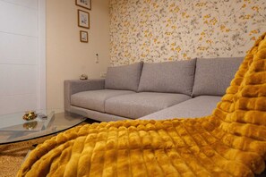 Ein Wohnzimmer mit grauem Sofa, einem Glastisch und einer gelben Decke.