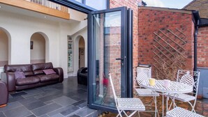 An open patio with a glass door, a brown leather sofa, and white metal chairs.