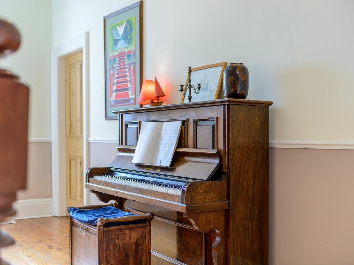 Un piano à queue se trouve dans une pièce où l’on aperçoit un banc en bois, un tableau encadré accroché au mur et une lampe posée sur le piano.